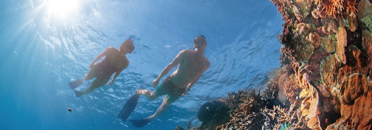 Two snorkelers swim in clear blue water near a colorful coral reef, with sunlight shining through the water's surface, illuminating the underwater scene. Various marine life and vibrant coral formations surround the snorkelers, making it one of the unique things to do in Bundaberg.