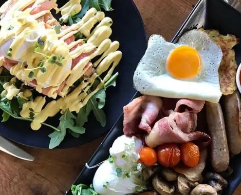 A hearty breakfast spread featuring a salad with greens and dressing on a black plate, and a platter with a sunny-side-up egg, bacon, sausages, roasted tomatoes, mushrooms, and a poached egg on a wooden table—perfect for fueling up before embarking on Bundaberg Tours to discover the unique things to do in Bundaberg.
