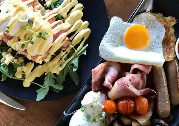 A hearty breakfast spread featuring a salad with greens and dressing on a black plate, and a platter with a sunny-side-up egg, bacon, sausages, roasted tomatoes, mushrooms, and a poached egg on a wooden table—perfect for fueling up before embarking on Bundaberg Tours to discover the unique things to do in Bundaberg.