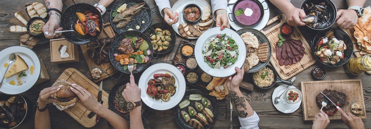 A top view of a crowded dining table with multiple people sharing a meal, reminiscent of the communal dining experiences you might find as one of the things to do in Bundaberg. Various dishes are spread across the table, including salads, meats, sandwiches, and desserts. Diners' hands are seen reaching for food, creating a lively and communal dining scene.