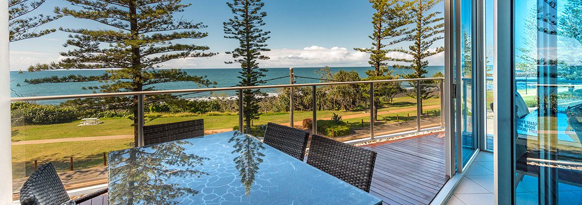 A glass table with wicker chairs on a spacious balcony overlooks a scenic ocean view—perfect for planning things to do in Bundaberg this weekend. Tall pine trees line the grassy area between the balcony and the beach, and the sky is clear with a few clouds. The balcony has glass railings and is attached to a modern building.