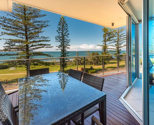 A glass table with wicker chairs on a spacious balcony overlooks a scenic ocean view—perfect for planning things to do in Bundaberg this weekend. Tall pine trees line the grassy area between the balcony and the beach, and the sky is clear with a few clouds. The balcony has glass railings and is attached to a modern building.