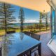 A glass table with wicker chairs on a spacious balcony overlooks a scenic ocean view—perfect for planning things to do in Bundaberg this weekend. Tall pine trees line the grassy area between the balcony and the beach, and the sky is clear with a few clouds. The balcony has glass railings and is attached to a modern building.
