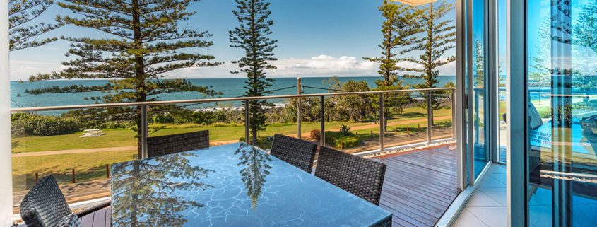 A glass table with wicker chairs on a spacious balcony overlooks a scenic ocean view—perfect for planning things to do in Bundaberg this weekend. Tall pine trees line the grassy area between the balcony and the beach, and the sky is clear with a few clouds. The balcony has glass railings and is attached to a modern building.
