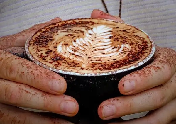 Close-up of a person holding a black cup of cappuccino or latte with intricate leaf latte art on the foamed milk surface. The hands, dusted with cocoa powder, suggest a cozy and artistic coffee moment—a perfect break during your Bundaberg Tours while exploring Unique Things To Do in Bundaberg. The background shows textured, light-colored fabric.