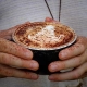 Close-up of a person holding a black cup of cappuccino or latte with intricate leaf latte art on the foamed milk surface. The hands, dusted with cocoa powder, suggest a cozy and artistic coffee moment—a perfect break during your Bundaberg Tours while exploring Unique Things To Do in Bundaberg. The background shows textured, light-colored fabric.