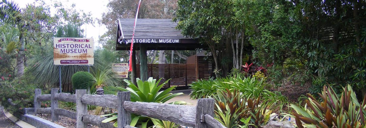 A rustic, wooden fence borders a path leading to the entrance of the Historical Museum, surrounded by lush green plants and trees. A sign to the left reads "Historical Museum" with additional text below it that is not fully readable—a must for Bundaberg Day Tours on your list of Things To Do Bundaberg.