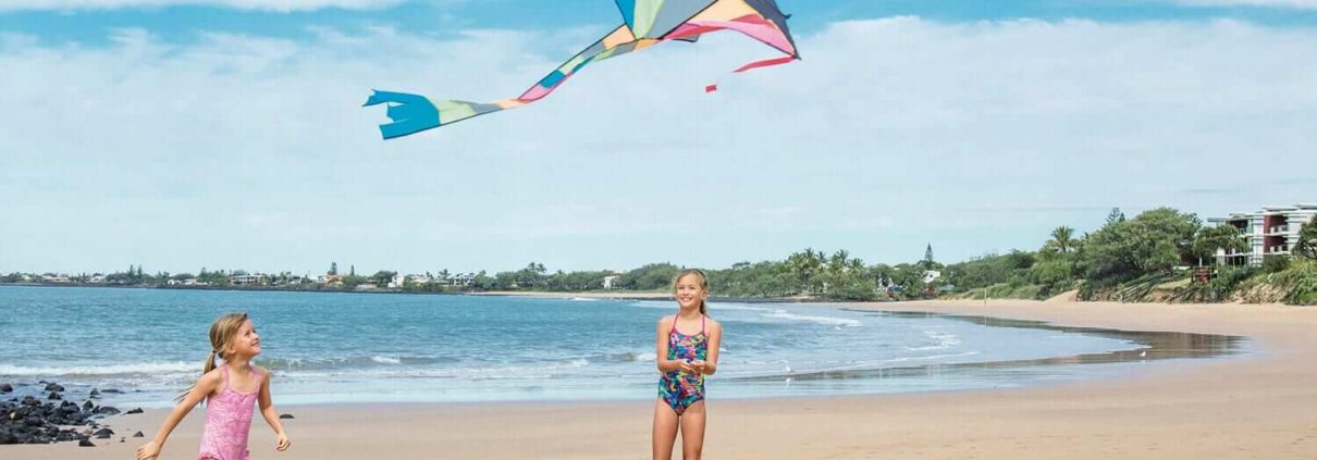 Two children in swimsuits are on a sandy beach under a clear blue sky, flying a colorful kite with long tails. The beach is expansive with gentle waves, and there's greenery and buildings in the background. With so many things to do in Bundaberg, they appear to be enjoying their time outdoors.