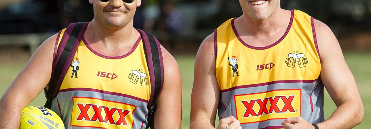 Two men wearing sleeveless sports jerseys with "XXXX" branding are smiling at the camera, enjoying one of the many Things To Do in Bundaberg. The man on the left wears sunglasses, a yellow cap, and a backpack, holding a yellow sports ball. The man on the right wears a burgundy cap and clenches his fists enthusiastically.