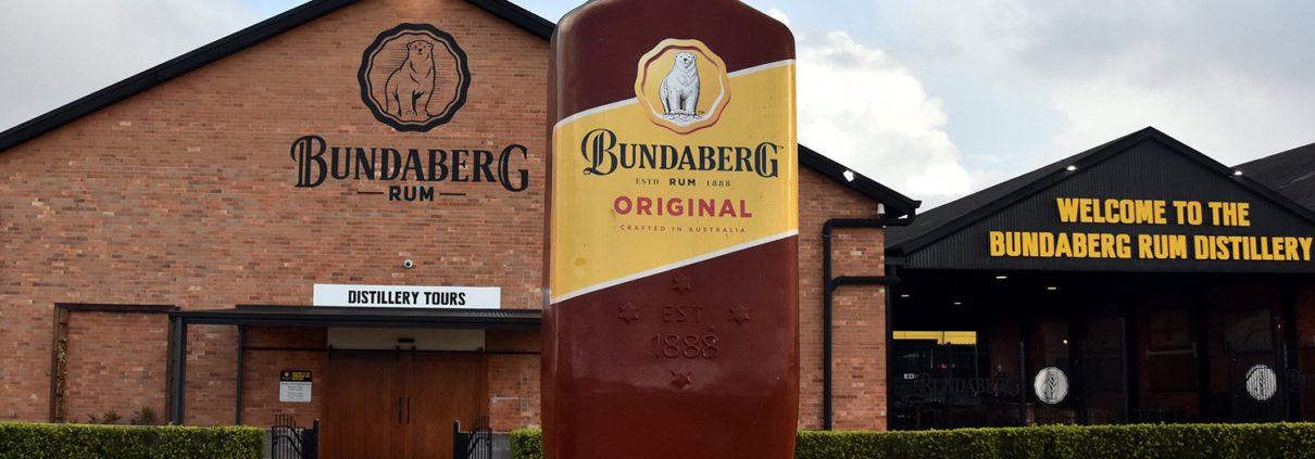 Exterior of the Bundaberg Rum Distillery features a large replica bottle of Bundaberg Original Rum out front. The distillery building boasts signs for "Distillery Tours" and "Welcome to the Bundaberg Rum Distillery." Perfect for things to do in Bundaberg, especially on one of the many Bundaberg day tours. The sky is partly cloudy.