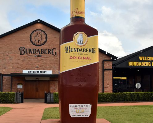 Exterior of the Bundaberg Rum Distillery features a large replica bottle of Bundaberg Original Rum out front. The distillery building boasts signs for "Distillery Tours" and "Welcome to the Bundaberg Rum Distillery." Perfect for things to do in Bundaberg, especially on one of the many Bundaberg day tours. The sky is partly cloudy.