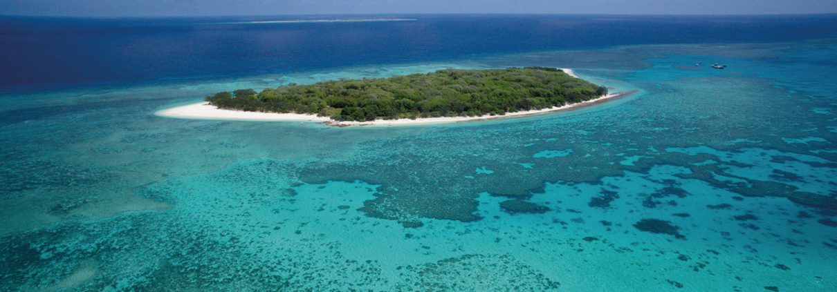 Aerial view of a small, lush green island surrounded by a white sandy beach and vibrant, turquoise blue waters with visible coral reefs. The horizon features a calm, expansive ocean under a partly cloudy sky — an ideal spot for Bundaberg Day Tours and exploring the many Things To Do Bundaberg has to offer.