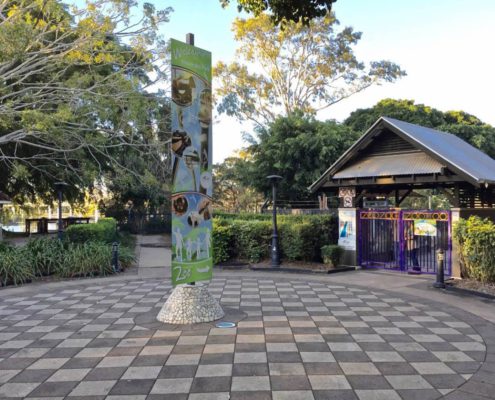 A spacious, checkered plaza with a tall, illustrated welcome sign in the center. In the background, an entrance gate to a zoo is indicated by colorful "ZOO" letters above the gate. Trees and foliage surround the area, providing a lush, green backdrop—perfect for exploring the top Things To Do Bundaberg.