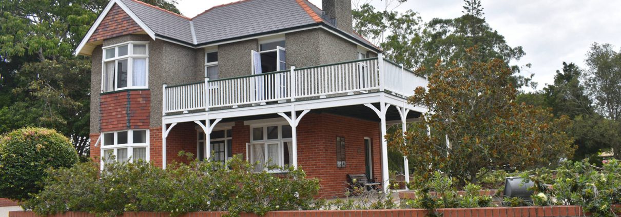 A two-story house features a mix of red brick and grey stucco exterior, white trim, and a wrap-around balcony on the second floor. The house is surrounded by a brick wall and lush greenery, with a neatly manicured lawn in front. Trees and shrubs provide ample shade—perfect for relaxing after exploring things to do in Bundaberg.