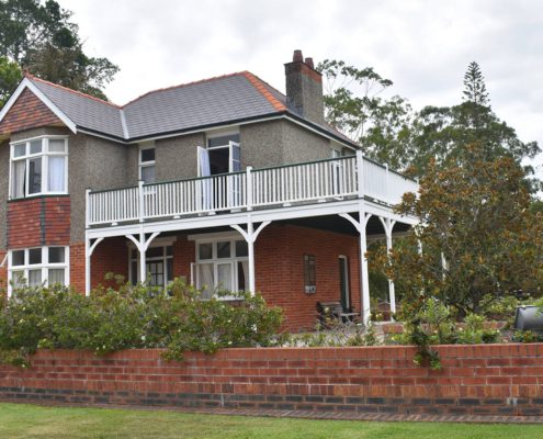 A two-story house features a mix of red brick and grey stucco exterior, white trim, and a wrap-around balcony on the second floor. The house is surrounded by a brick wall and lush greenery, with a neatly manicured lawn in front. Trees and shrubs provide ample shade—perfect for relaxing after exploring things to do in Bundaberg.