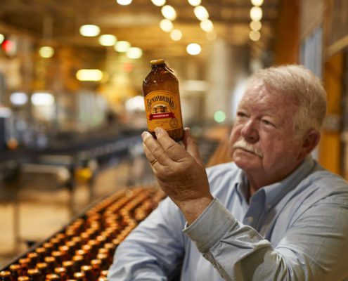 An older man with white hair and a mustache holds a bottle of Bundaberg Ginger Beer, examining it closely. He is standing in a brewery with rows of bottles stretching out behind him under warm lighting. The background is slightly blurred, focusing on the man and bottle—one of the top things to do in Bundaberg.