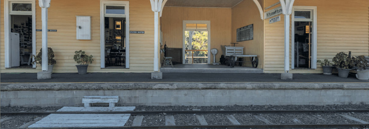 A rustic yellow railway station building with a sign reading "Bundaberg Railway Museum" invites you to explore one of the top things to do in Bundaberg. The platform features benches, potted plants, and doors marked "Ladies Waiting Room" and "Station Master's Office," with railway tracks and trees in the background.