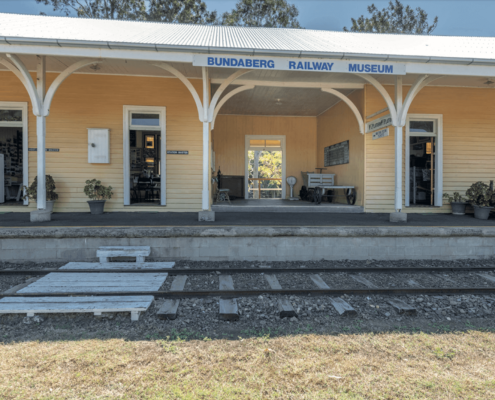 A rustic yellow railway station building with a sign reading "Bundaberg Railway Museum" invites you to explore one of the top things to do in Bundaberg. The platform features benches, potted plants, and doors marked "Ladies Waiting Room" and "Station Master's Office," with railway tracks and trees in the background.